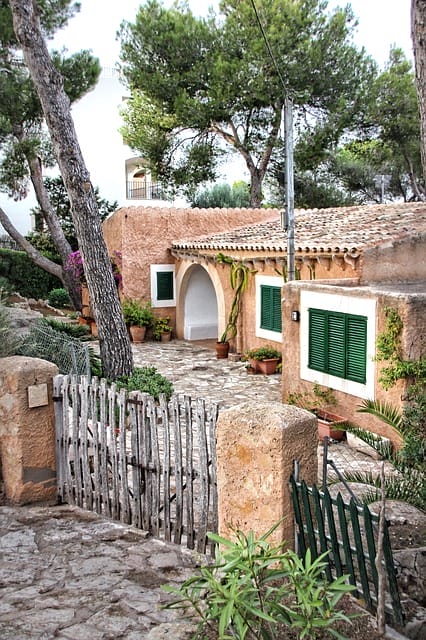 Verlassene Mini-Finca auf Mallorca mit Holzzaun und grünen Fensterläden, umgeben von Bäumen