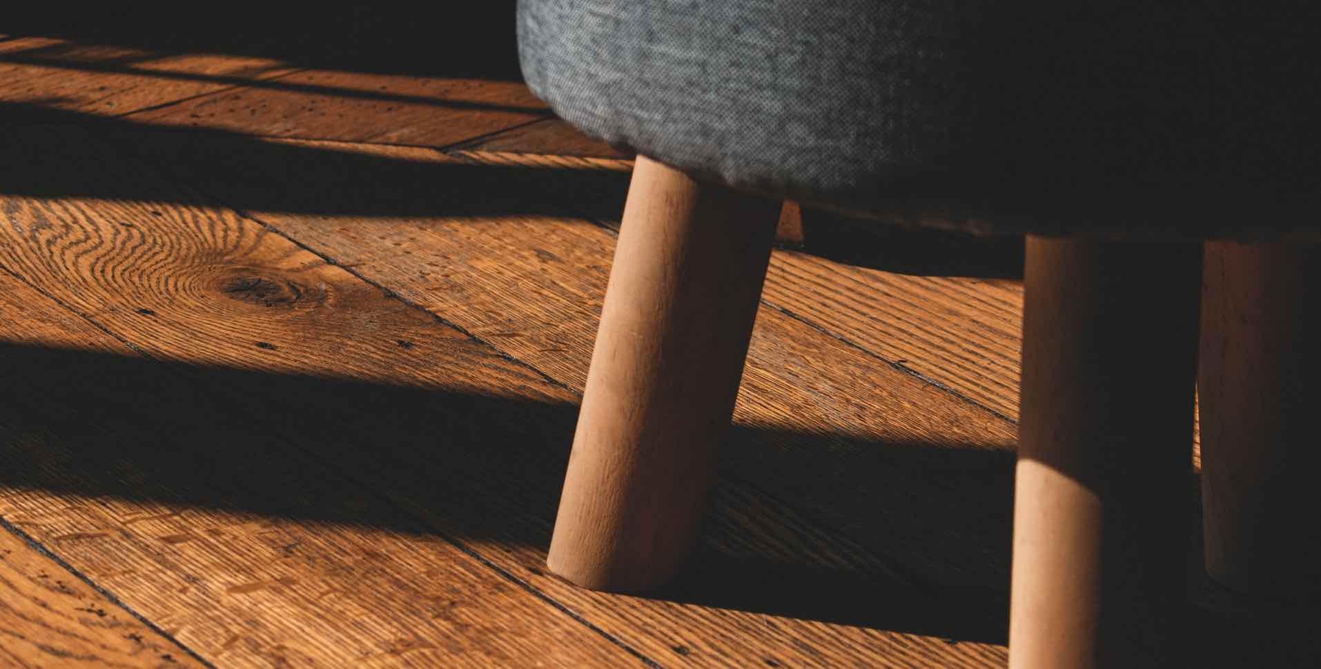 Holzfußboden mit Sonnenschatten und dem Bein eines Hockers mit grauem Sitzpolst stoffbezogenem Sitz
