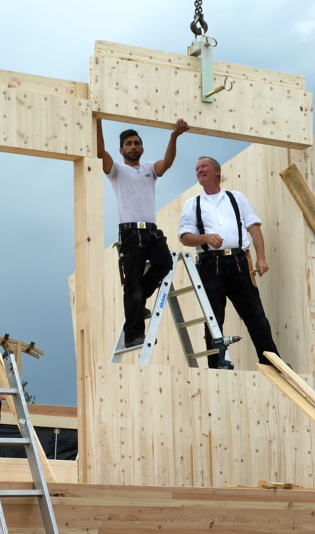 Zwei Bauarbeiter in traditioneller Kleidung errichten das Holzgerüst eines Hauses, der linke Mann steht auf einer Krause Leit