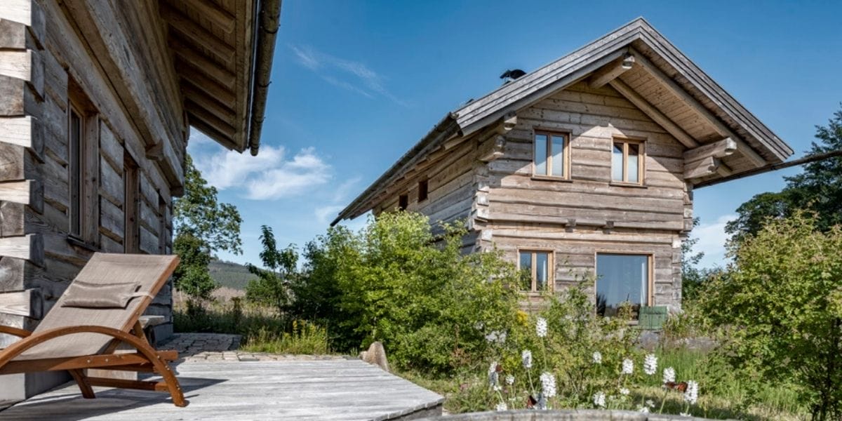 Zwei Blockhäuser aus Naturstämmen mit Holzdach und Veranda, umgeben von grünen Büschen und weiß blühenden Pflanzen