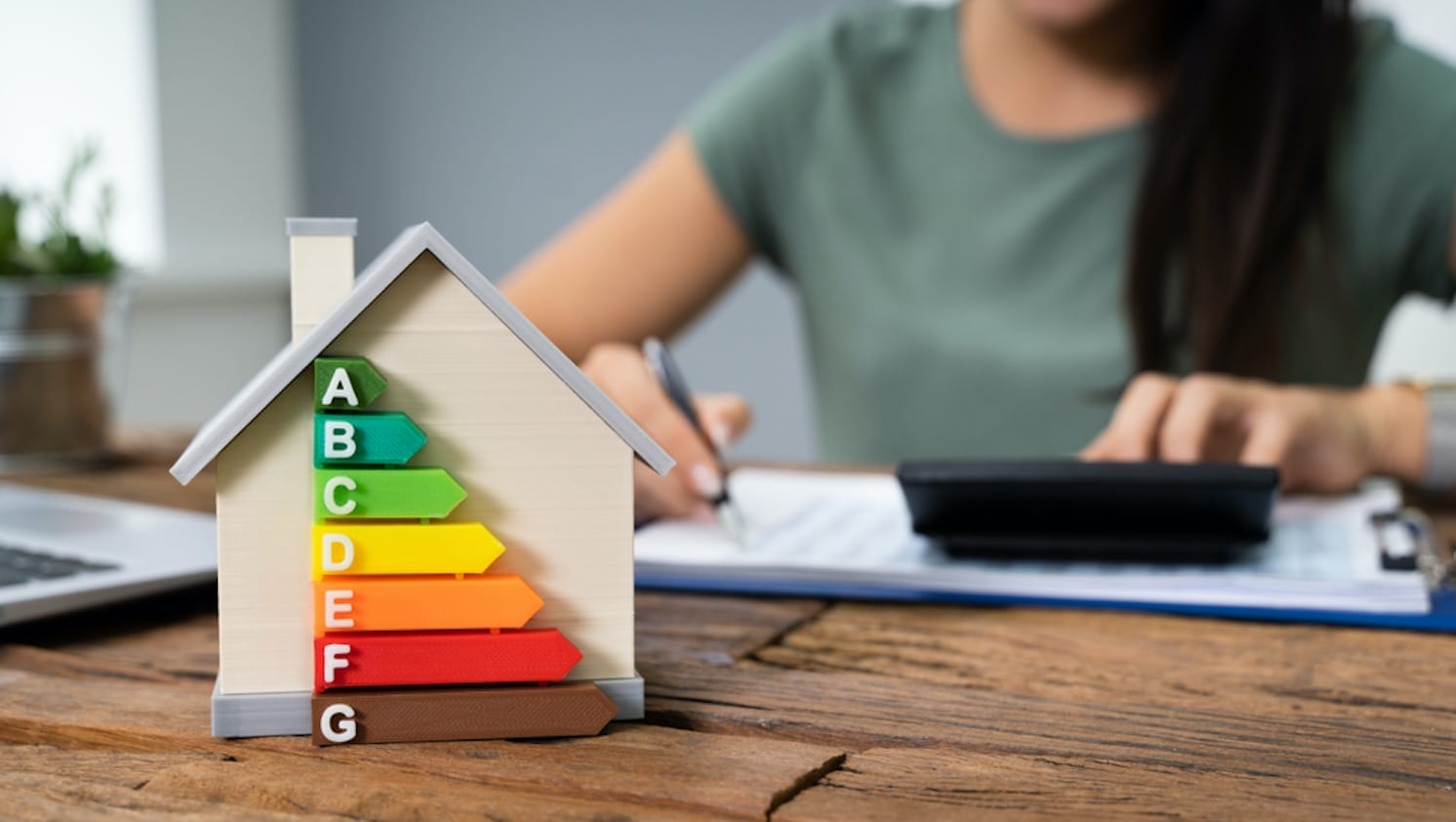Ein Modellhaus mit Energieeffizienz-Skala von A bis G, im Hintergrund ein Mensch, der Notizen macht und einen Taschenrechner