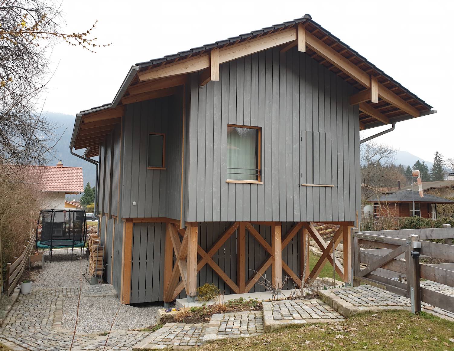 Modernes graues Holzhaus auf Holzbalken über einer Garage, Kopfsteinpflasterweg und kleiner Garten