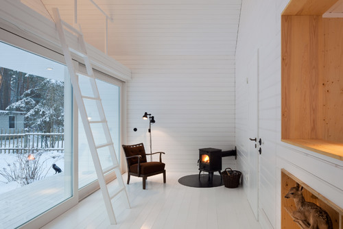 Heller Raum mit vielen Weißtönen, Kaminofen, braunen Sessel und Sprossenleiter vor einer großen Fensterfront mit Winterlandsc