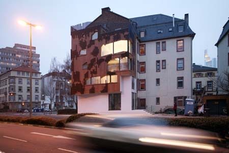 Rostbraunes, schmales Haus mit großen Fenstern in Baulücke integriert, davor eine Straße mit verschwommenem Auto