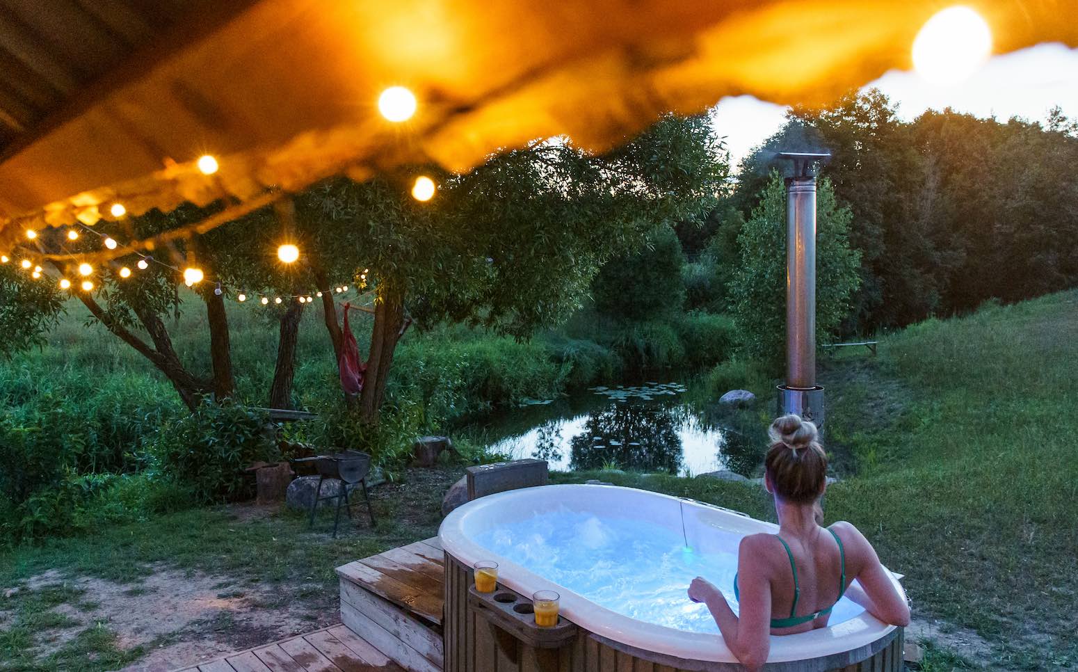 Frau im Whirlpool mit Blick auf einen kleinen Teich, umgeben von Bäumen, Lichterkette