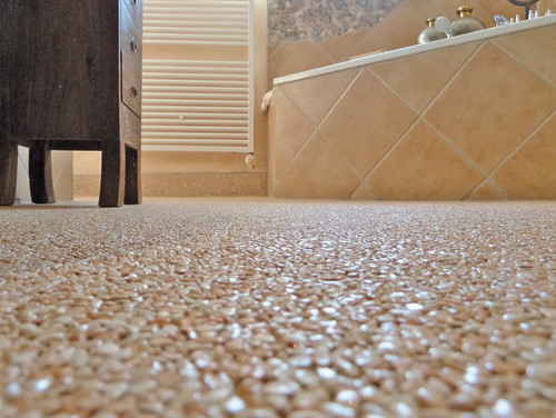 Nahaufnahme eines schimmernden, beigefarbenen Steinteppich-Bodens in einem Badezimmer mit Badewanne und Schrank