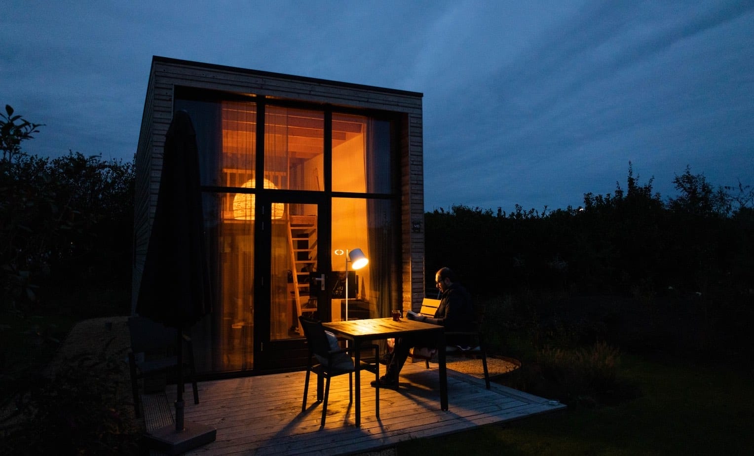 Moderner Mann liest auf einer Terrasse am Holztisch vor einem Minihaus bei Dämmerung