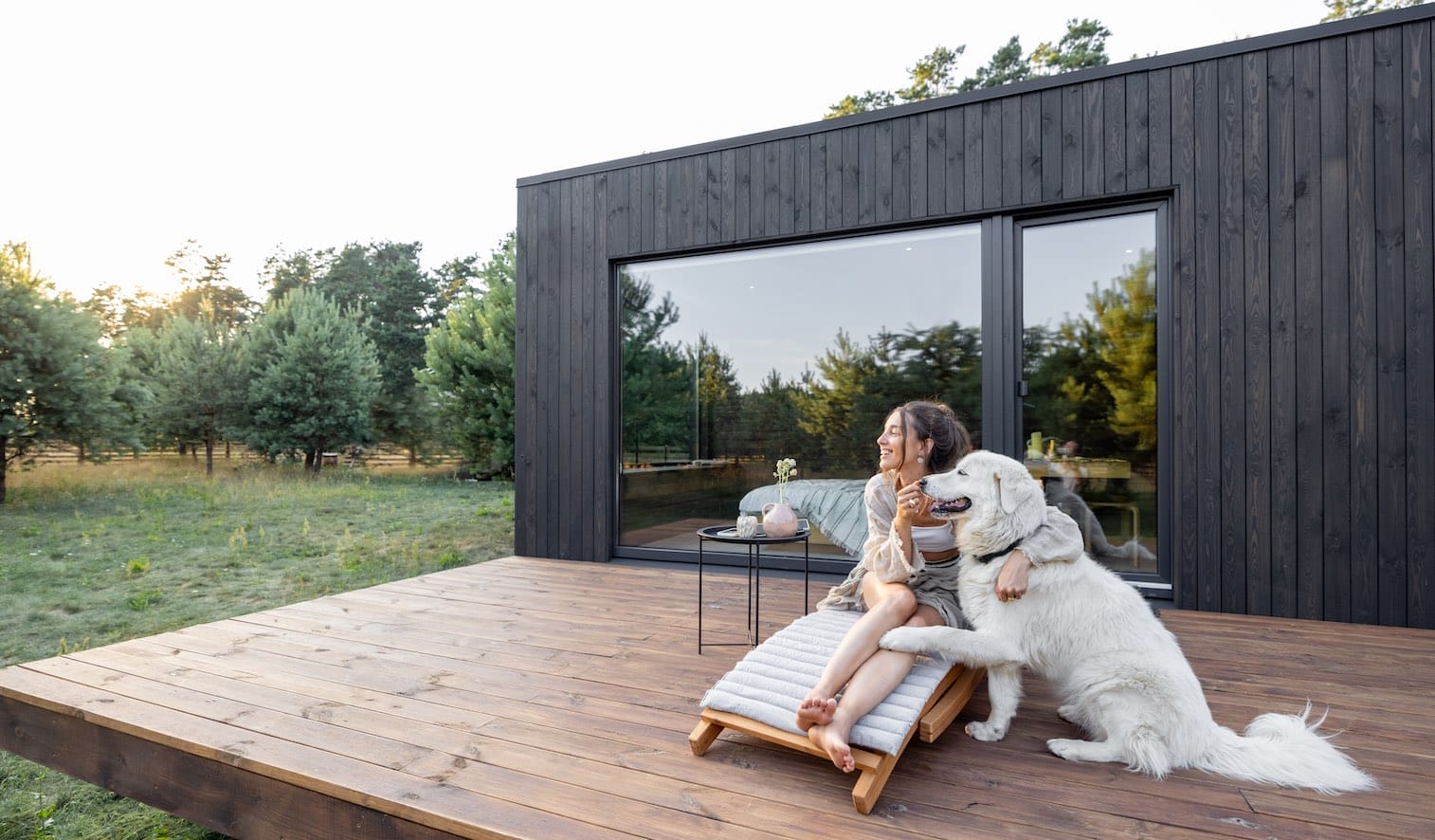Eine Frau und ein großer weißer Hund sitzen entspannt auf einer Holzterrasse vor einem modernen Haus