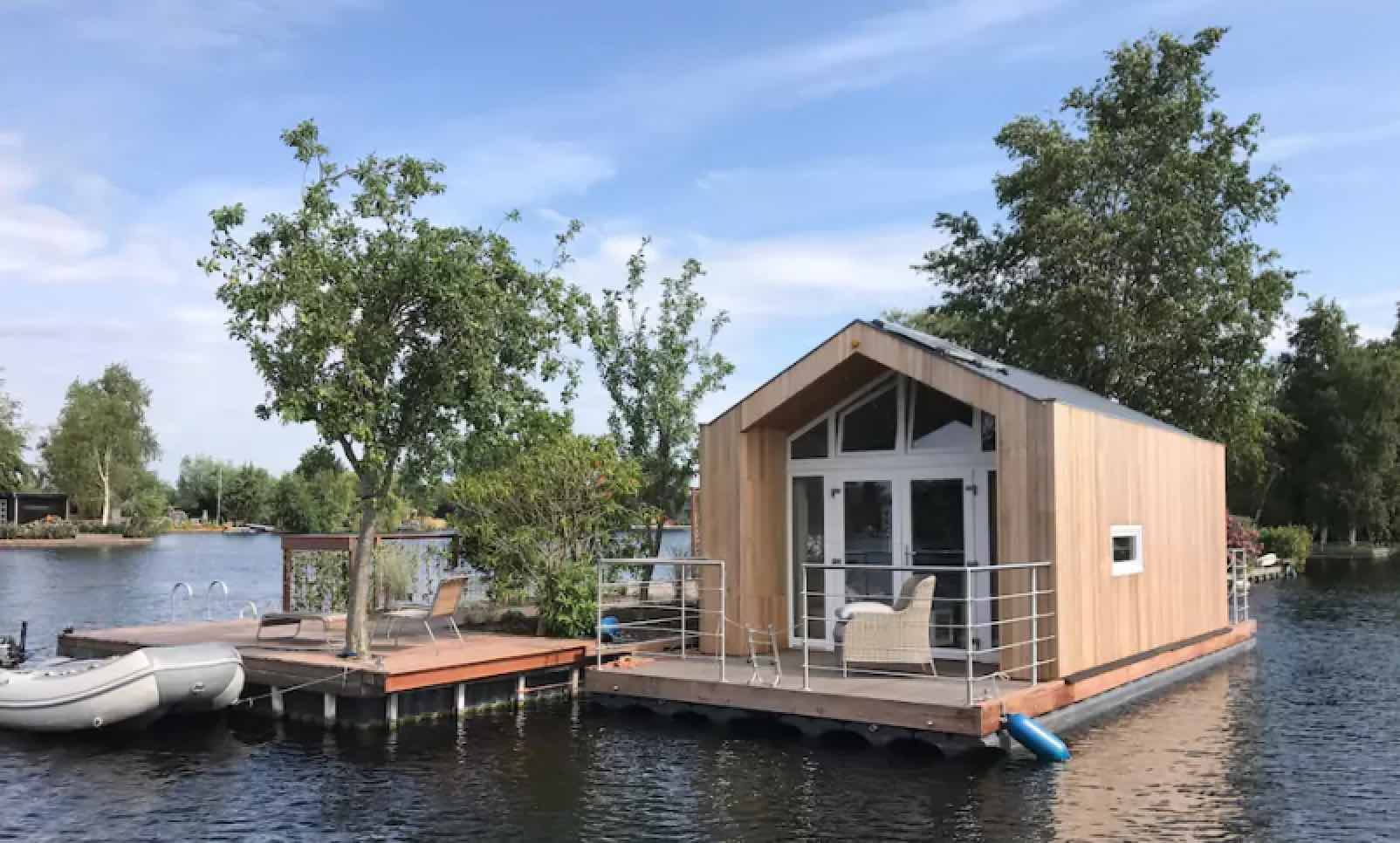 Modernes Holz-Hausboot mit Terrasse und kleinem Schlauchboot auf ruhigem Gewässer umgeben von Bäumen und Sträuchern