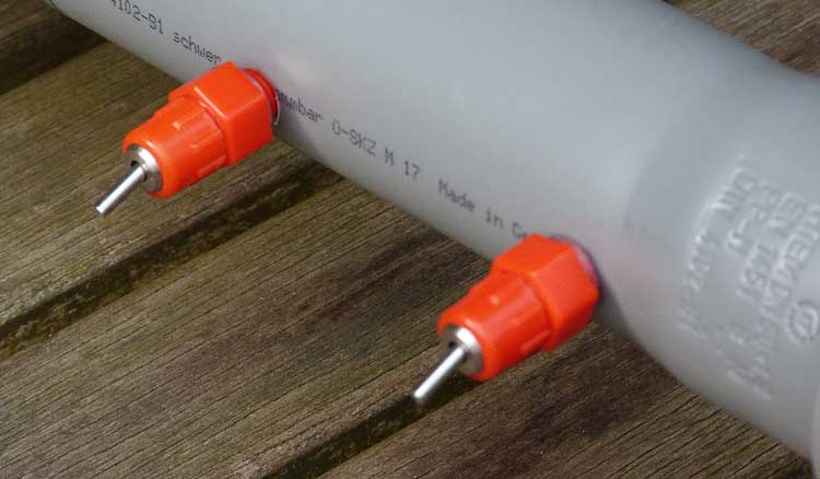Nahaufnahme eines grauen Abflussrohrs mit zwei orangen Nippeltränken auf Holzdielen, Made in Germany
