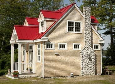Ein kleines Haus mit rot gedecktem Dach, Holzfassade und hohem Steinkamin am Waldrand