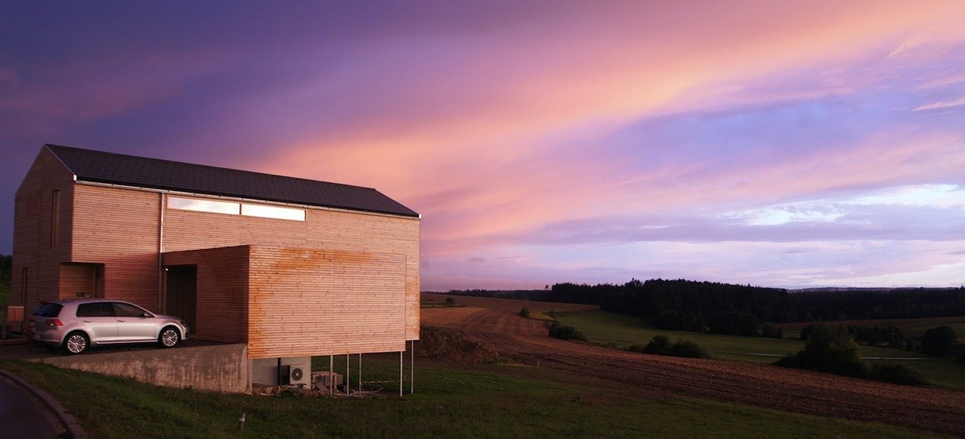 Modernes Modulhaus mit Holzfassade und schwarzem Dach, davor ein silberner VW Golf. Himmel mit pinken Wolken