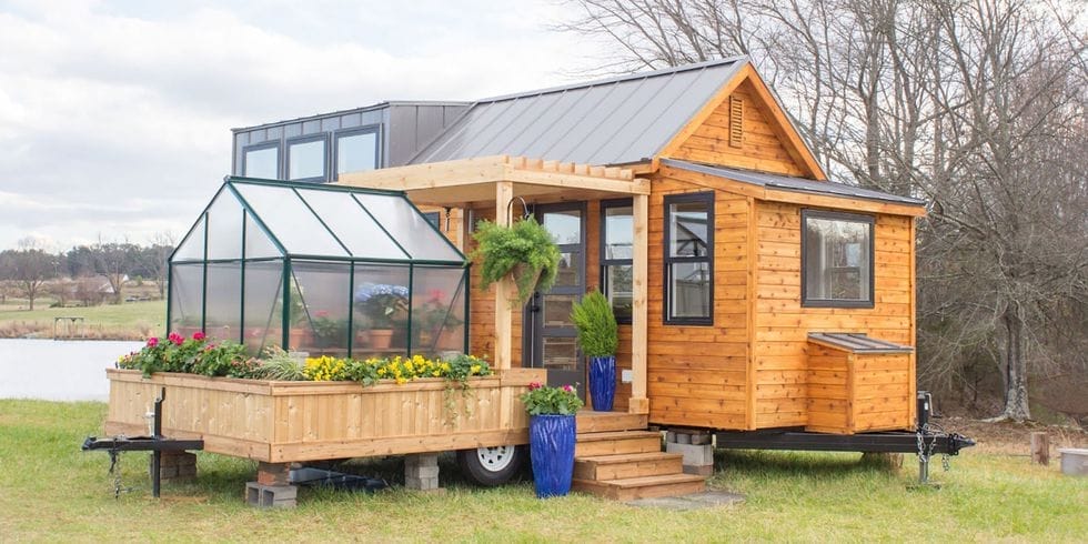 Ein kleines, holzverkleidetes Tiny House auf Rädern mit Veranda, Gewächshaus und Blumenkästen auf grünem Gras
