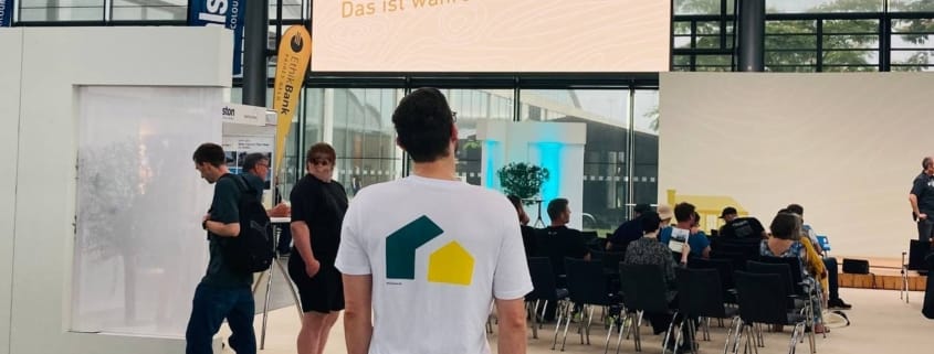 Ein Mann mit weißem T-Shirt und dem New Housing Logo steht in einer Messehalle mit Publikum und Leinwand
