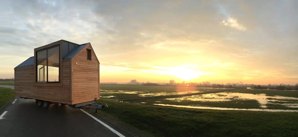 Ein Tiny House mit großen Fenstern an der Seite auf einem Anhänger stehend, im Hintergrund geht die Sonne unter