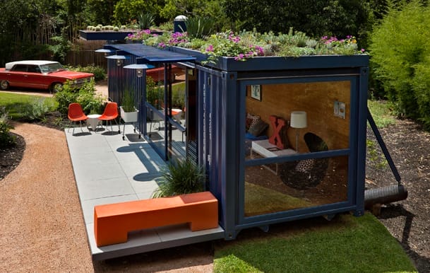 Ein umgebauter blauer Seecontainer mit begrünter Dachfläche, umgeben von einem Garten mit Terrasse