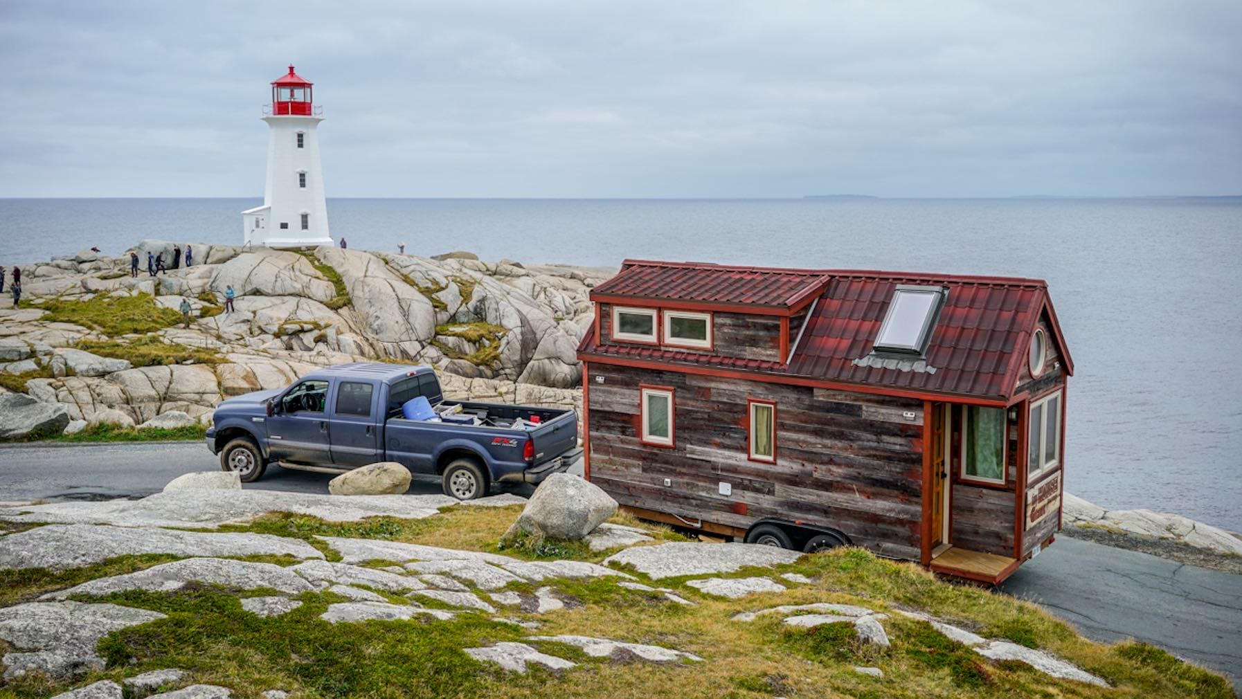 Ein Tiny House, gezogen von einem Pick-up-Truck, an der Küste mit einem Leuchtturm im Hintergrund