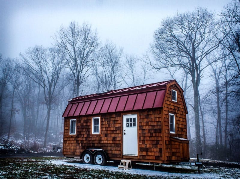 Ein Tiny House mit rot gedecktem Mansarddach auf einem Anhänger, umgeben von Bäumen und Schnee