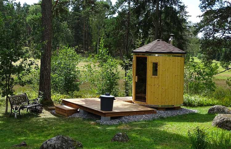 Runde Holz-Minisauna mit dunklem Dach und Kamin auf einer Holzterrasse im Grünen, umgeben von Bäumen