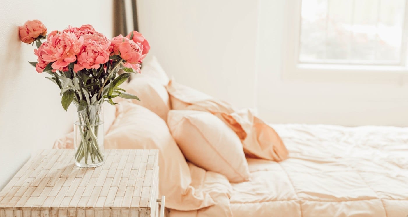 Rosen in Vase auf Nachttisch neben einem Bett mit Rosé-Bettwäsche und Fenster im Tiny House