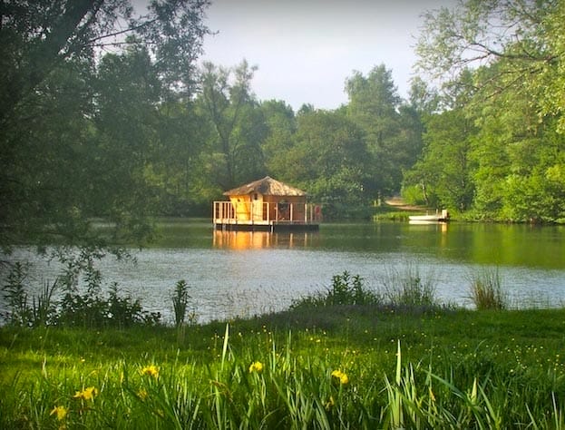 Ein schwimmendes Holz-Baumhaus mit Strohdach auf einem ruhigen See umgeben von viel Grün im Val de Bonnal
