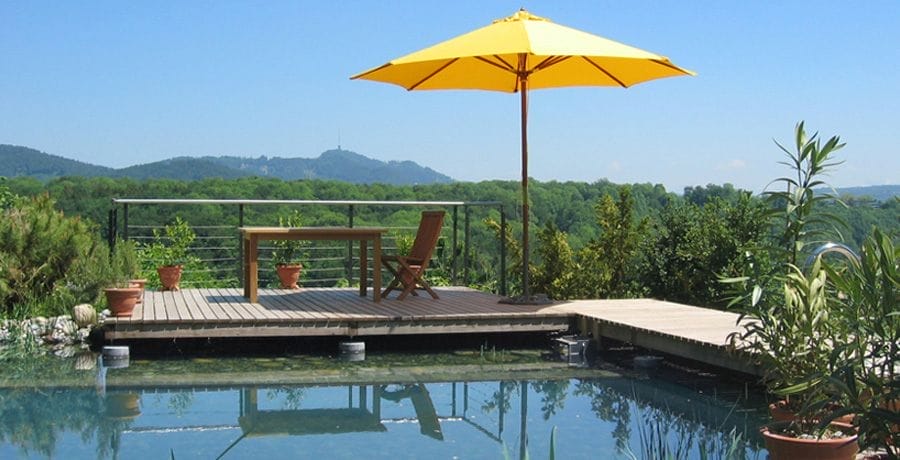 Holzterrasse am Naturpool mit gelbem Sonnenschirm, Tisch und Stühlen vor einer bewaldeten Hügellandschaft