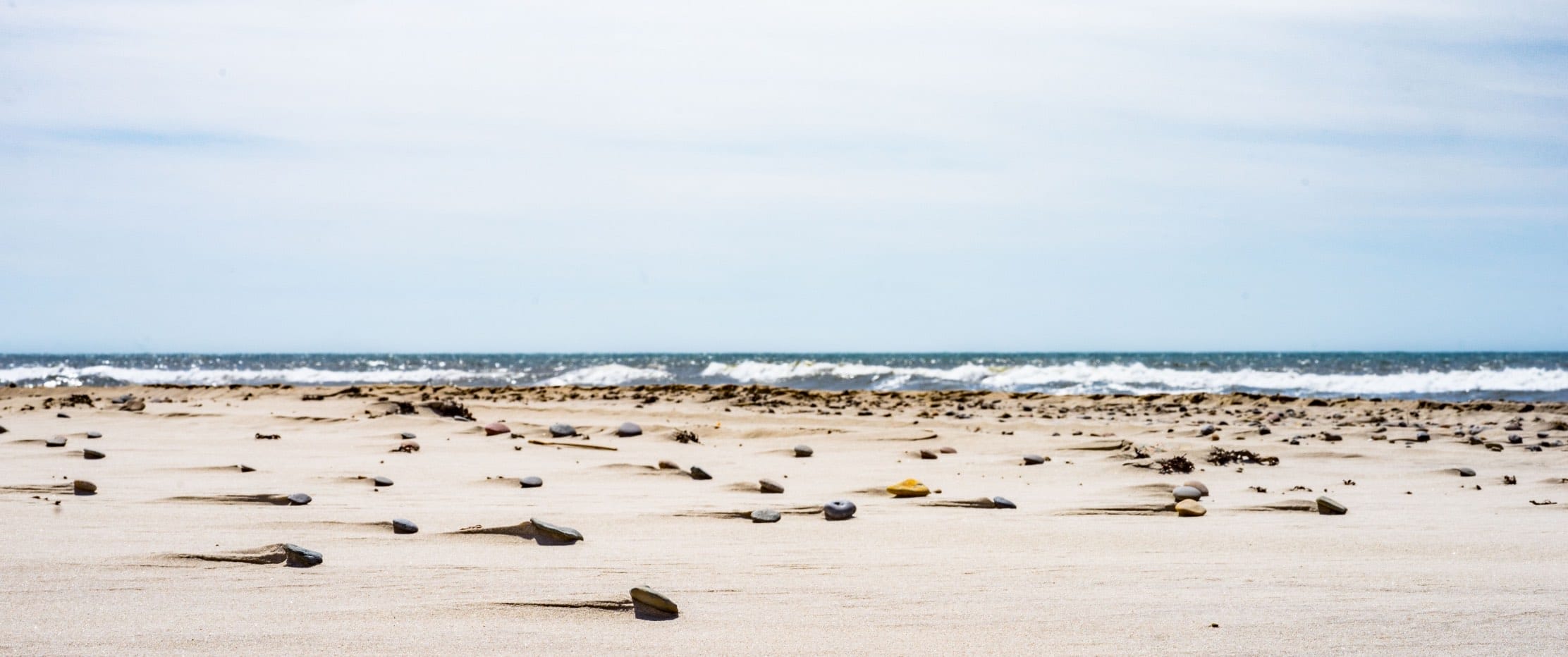 Sandstrand mit einzelnen Kieselsteinen und Meereswellen im Hintergrund