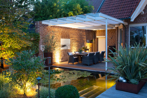 Blick von einem Garten mit Teich und angestrahlten Pflanzen auf eine überdachte Holzterrasse mit Esstisch und Stühlen