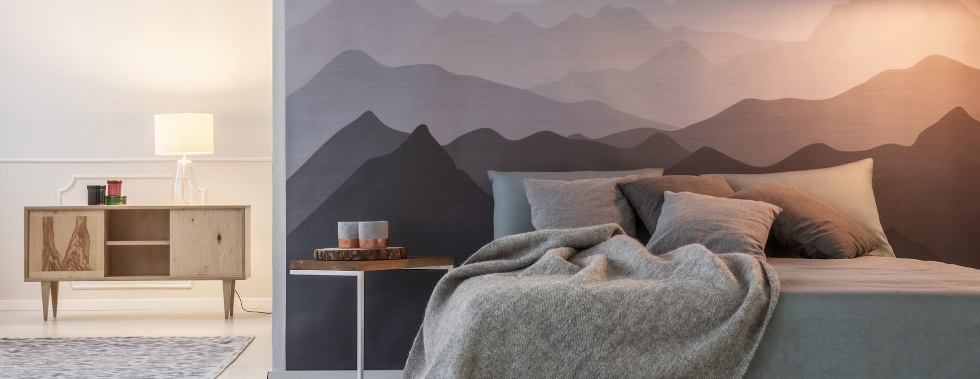 Schlafzimmerwand mit Bergtapete hinter hellgrauem Bett mit Kissen und Decke, links Sideboard mit Lampe