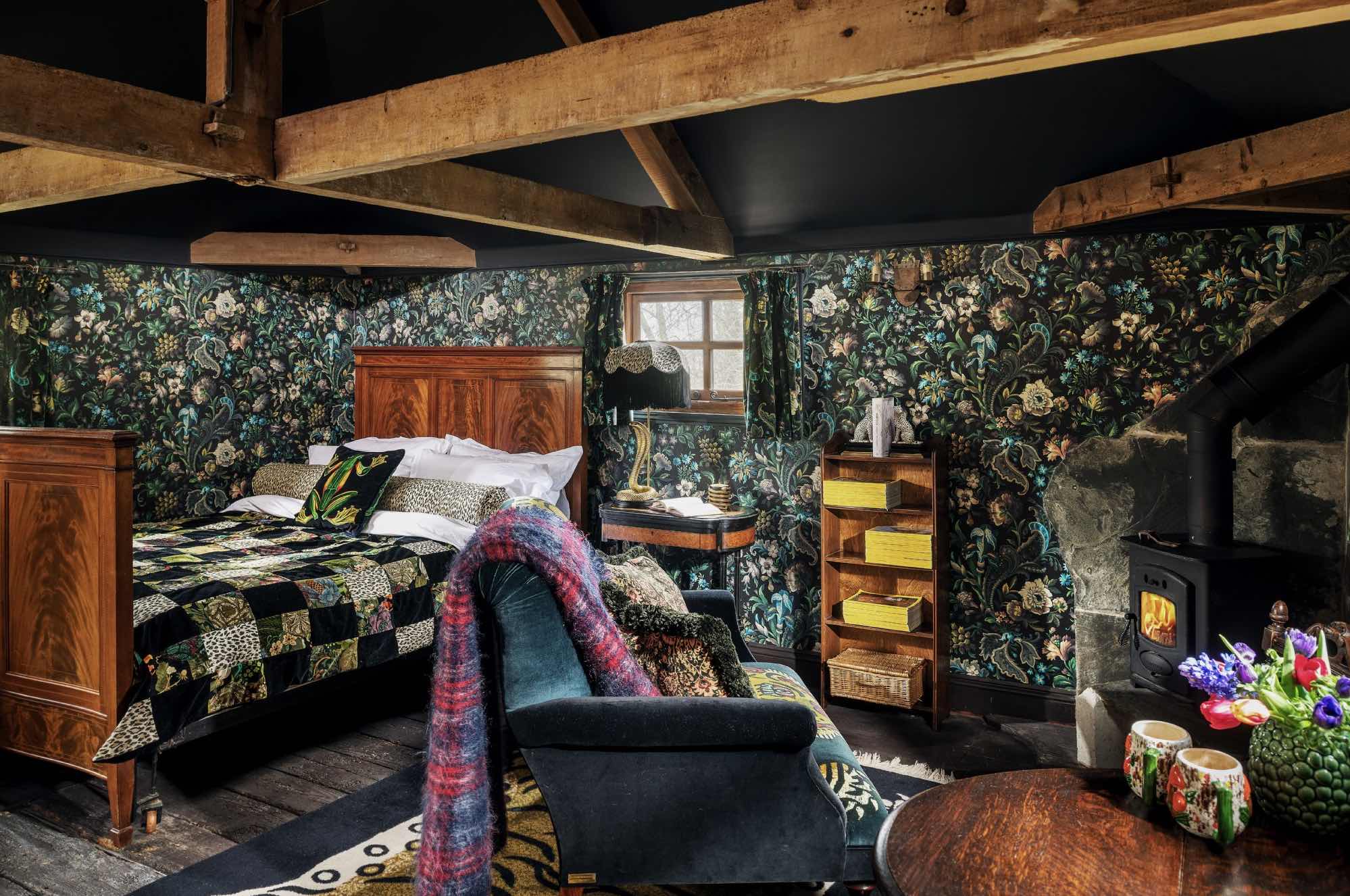 Ein dunkles Schlafzimmer mit gemusterter Blumentapete, Holzbett, Kaminofen und groben Deckenbalken an der Decke