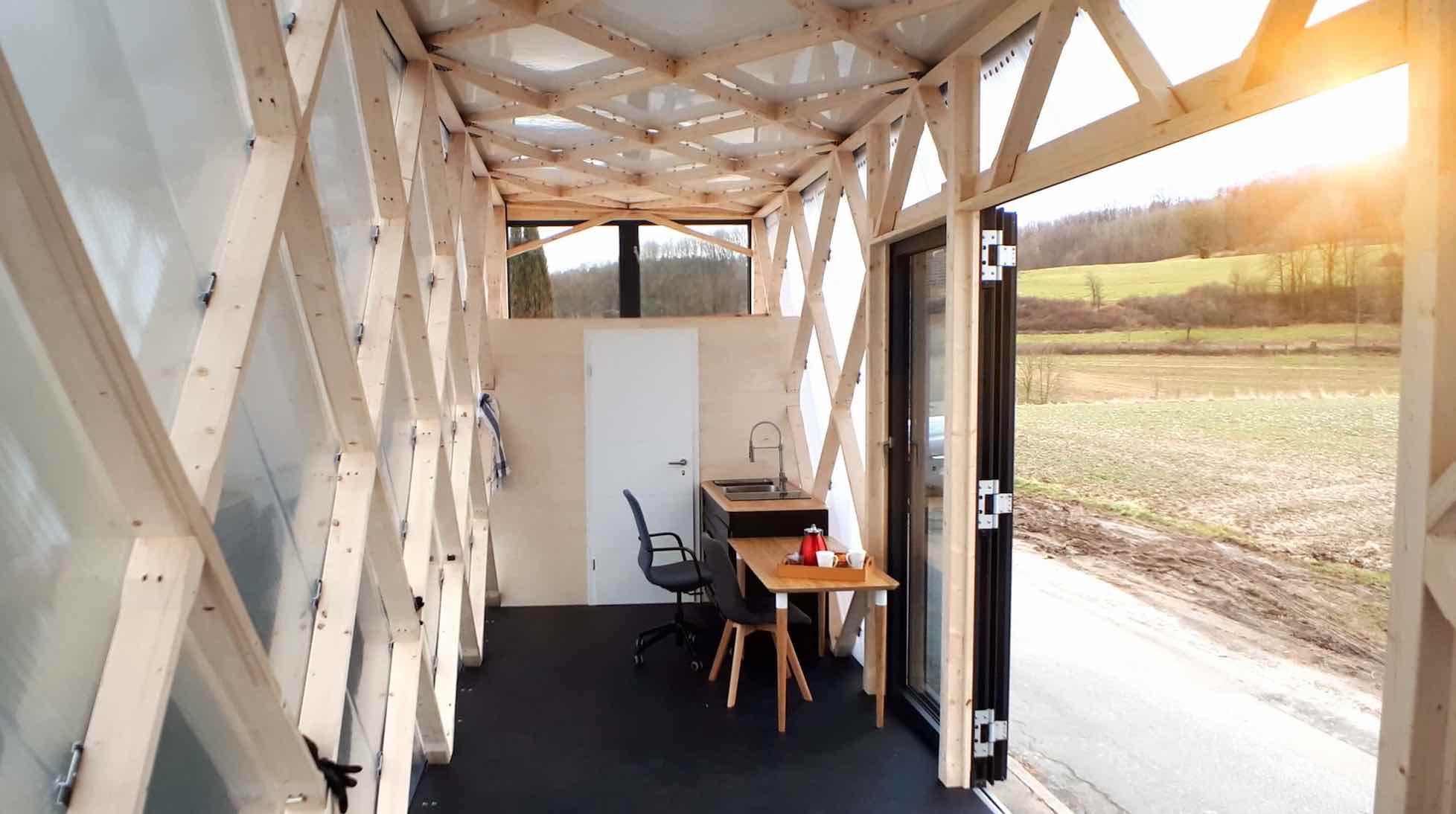 Innenraum eines Tiny House mit Holzbalkenkonstruktion, Teekessel auf Tisch und Blick ins Grüne