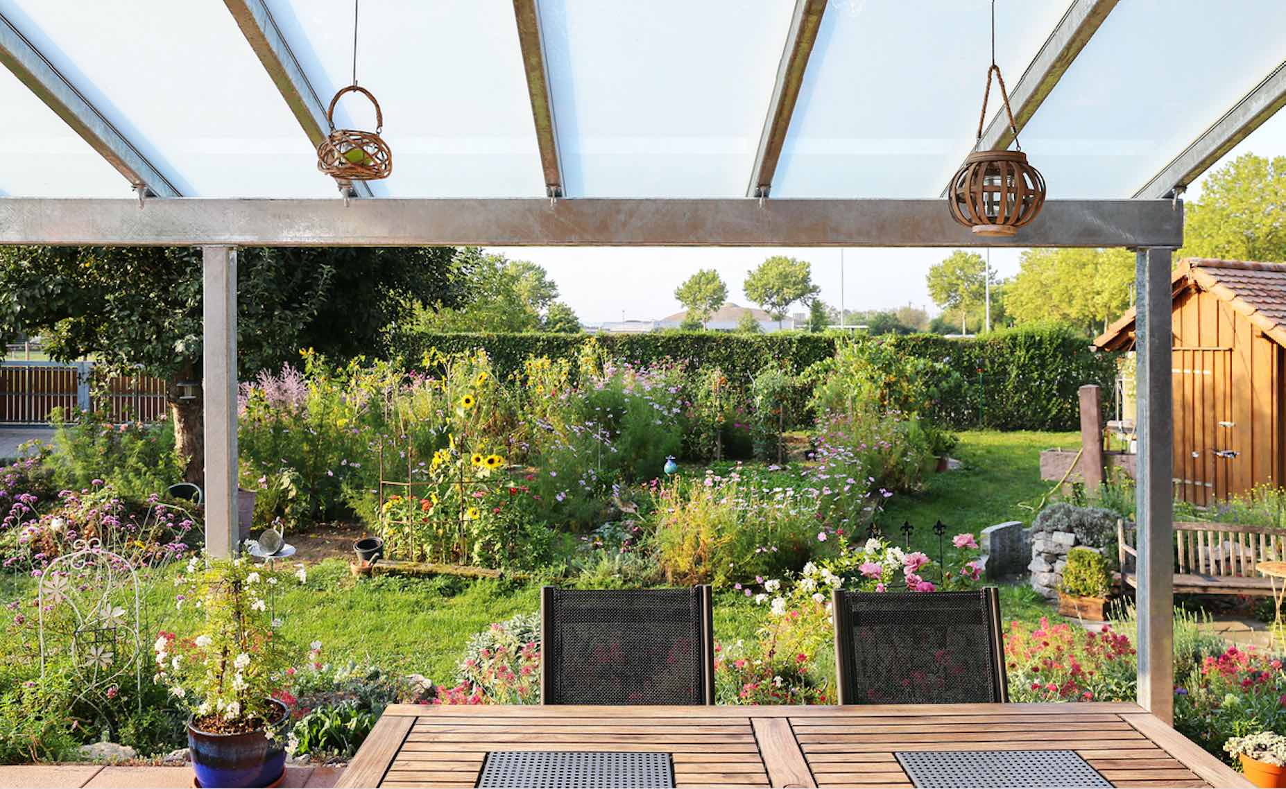 Blick von einer Terrassenüberdachung mit Holztisch und Gartenstühlen in einen blühenden Garten mit Rasenfläche