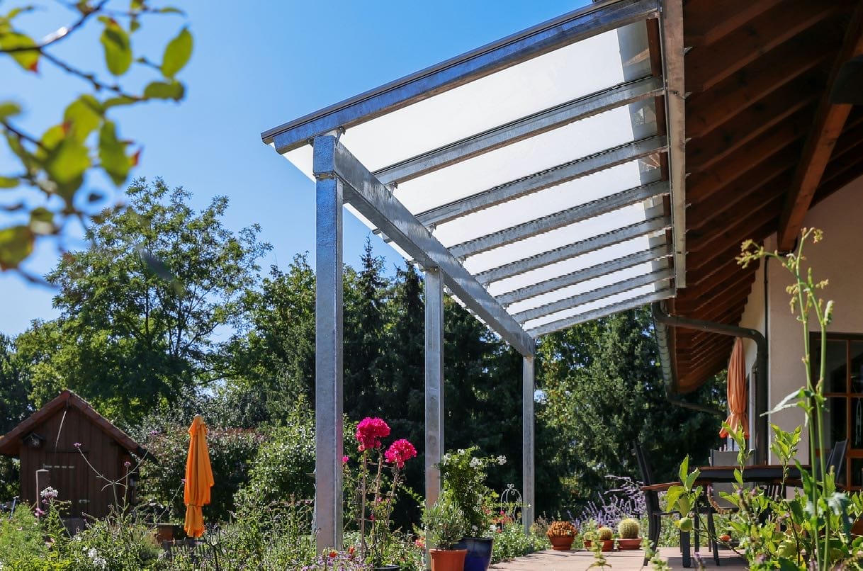 Stabile Terrassenüberdachung aus Leichtmetall vor ländlichem Hintergrund mit viel Grün und kleinen Holzhäuschen