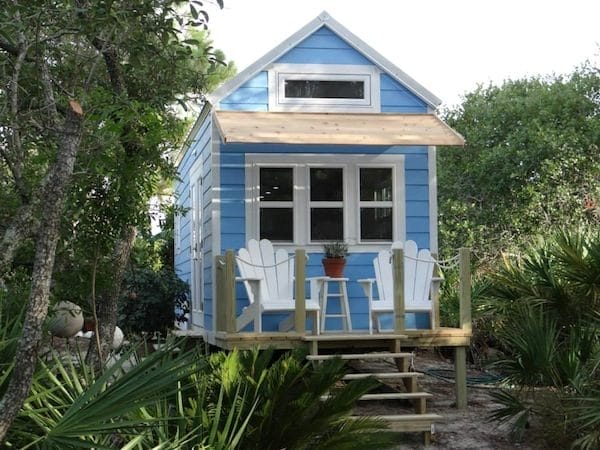 Blaues Mini-Haus umgeben von viel Grün mit einer kleinen Veranda und zwei weißen Stühlen davor