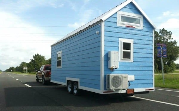 Blaues rollbares Tiny House mit Satteldach und Klimaanlage wird auf einer Straße von einem Pickup-Truck gezogen