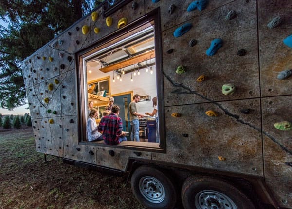 Seitenansicht eines Tiny House mit Kletterwand außen und offener Seite, in der Menschen an einem Tisch sitzen
