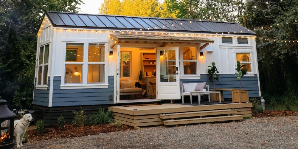 Modernes Tiny House mit weißer Fassade und blauem Sockel, Veranda und Lichterkette im Grünen. Ein Hund steht links