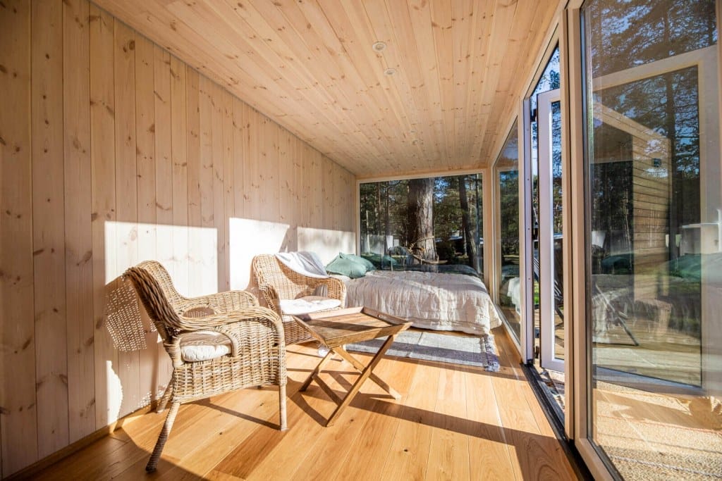 Innenraum eines PopUpHuts mit Holzboden und -wänden, dahinter ein Bett und großer Fensterfront zur Natur