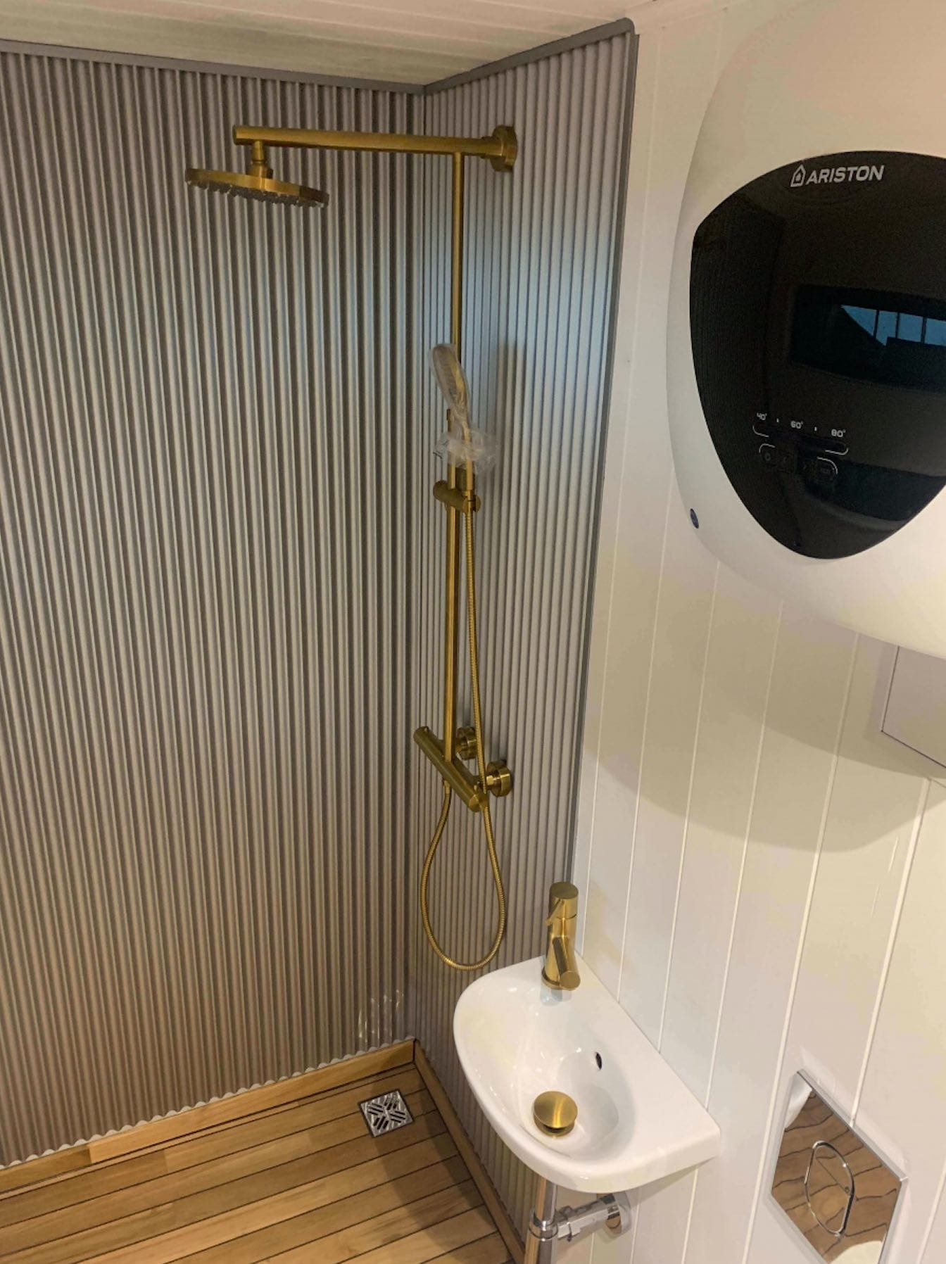 Badezimmer mit goldener Duscharmatur und kleiner Waschgelegenheit, daneben ein Warmwasserbereiter von Ariston