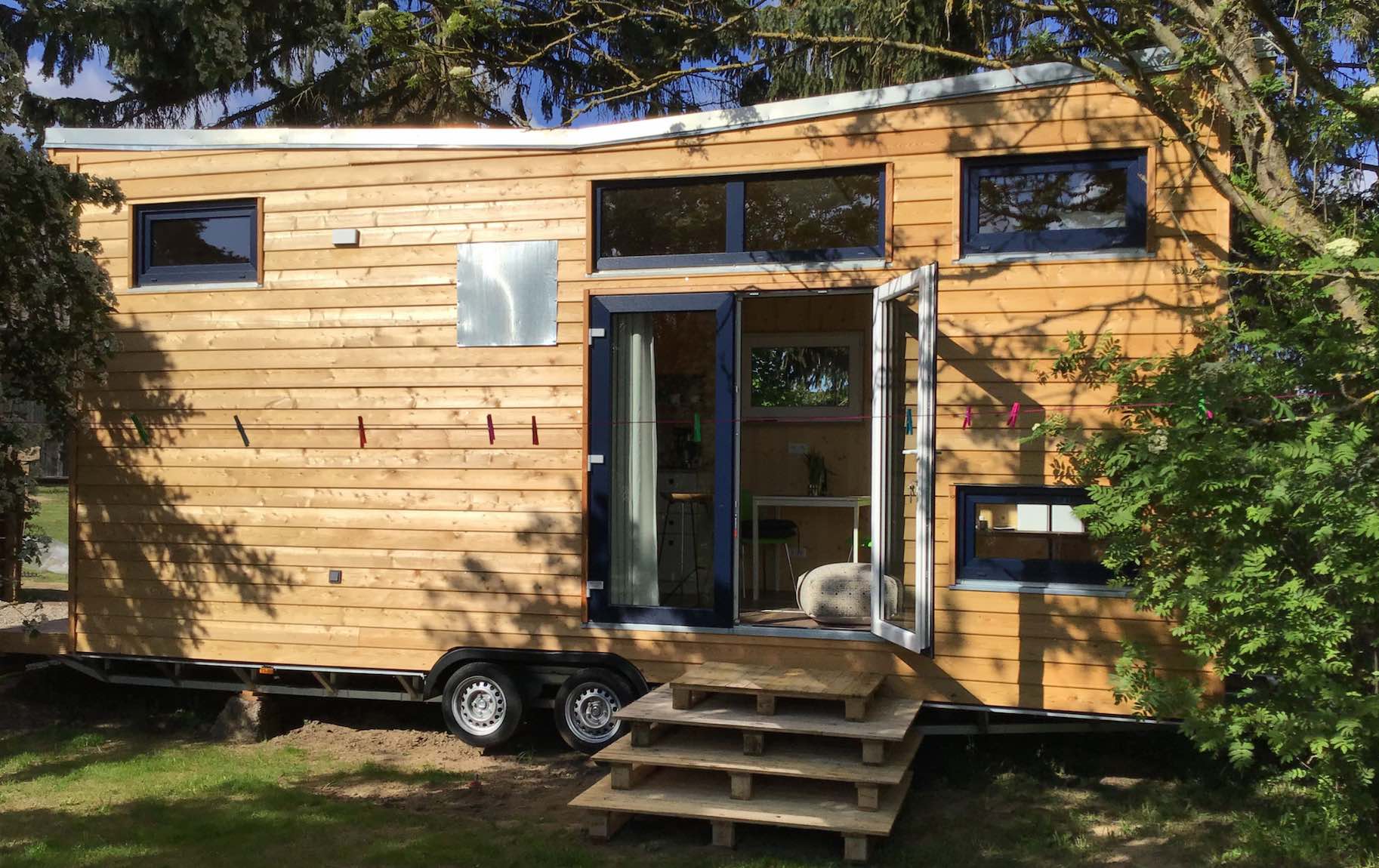 Holzverkleidetes Tiny House auf Rädern mit offener Tür, davor eine Holztreppe aus Palettenelementen