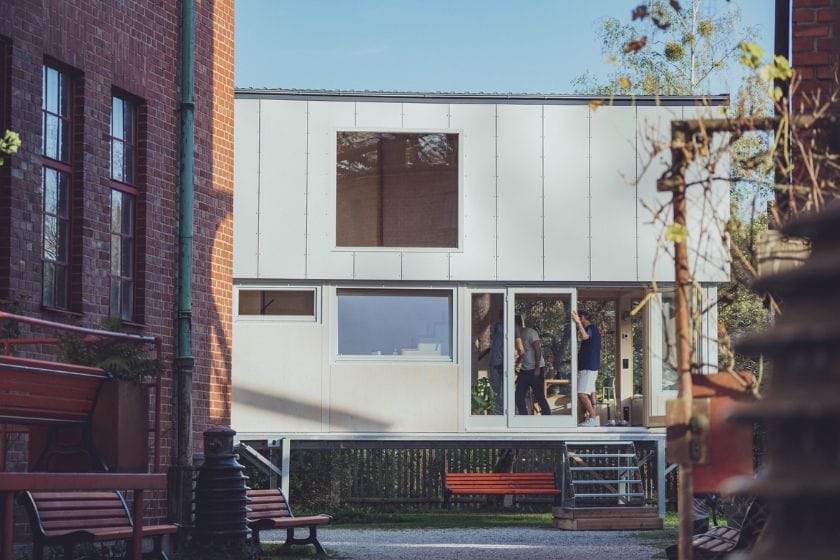 Ein Tiny House mit weißer Fassade und großen Fenstern steht auf einem Fundament neben einem Backsteingebäude