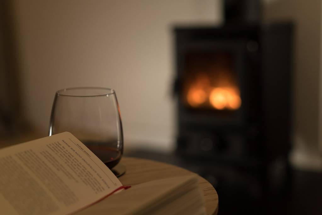 Ein Glas Wein und ein offenes Buch auf einem Holztisch vor dem brennenden Hobbit-Holzofen