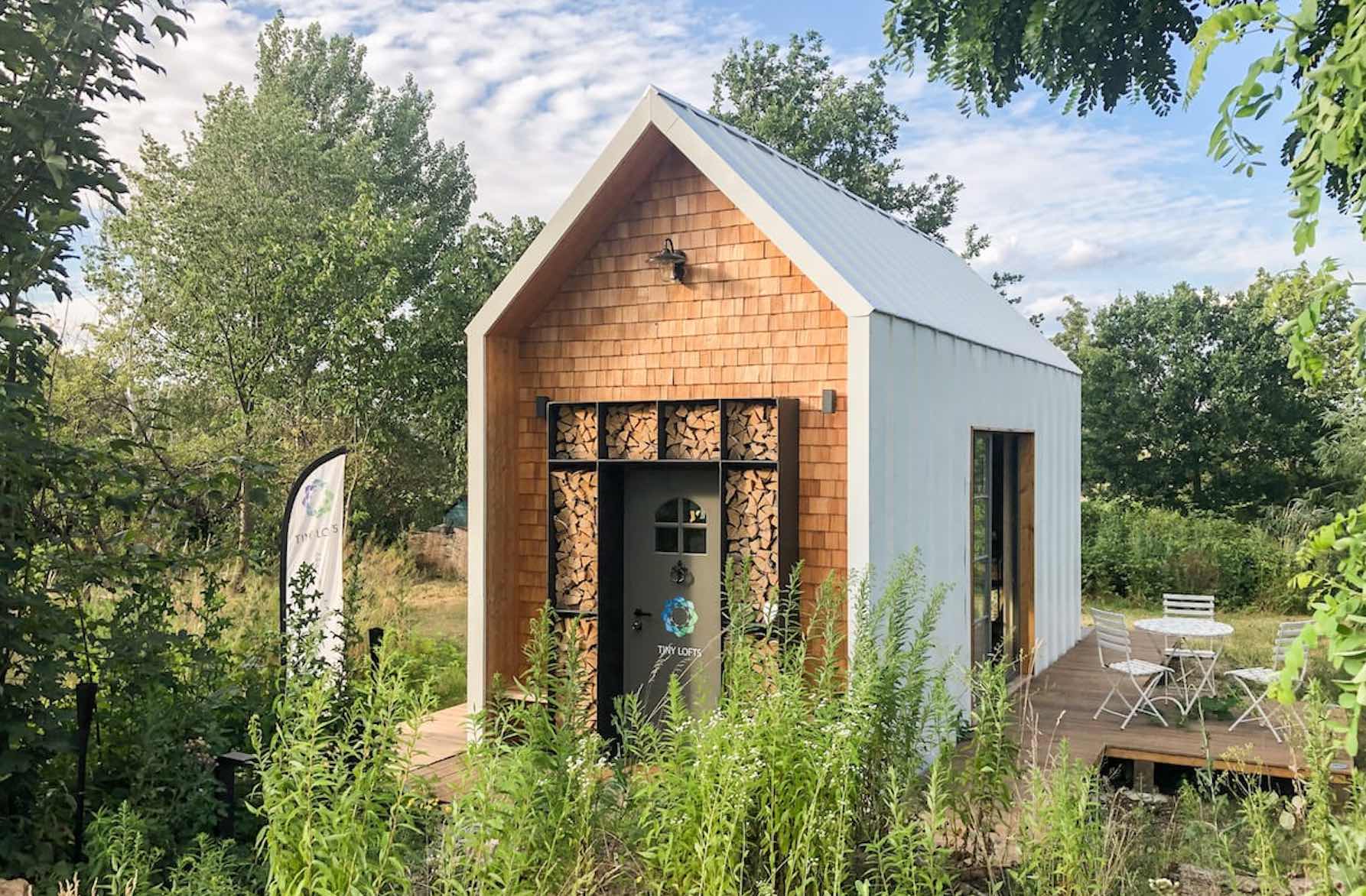 Ein Tiny House mit Holzfassade und hellem Dach, daneben eine kleine Terrasse mit Gartenmöbeln und viel Grün