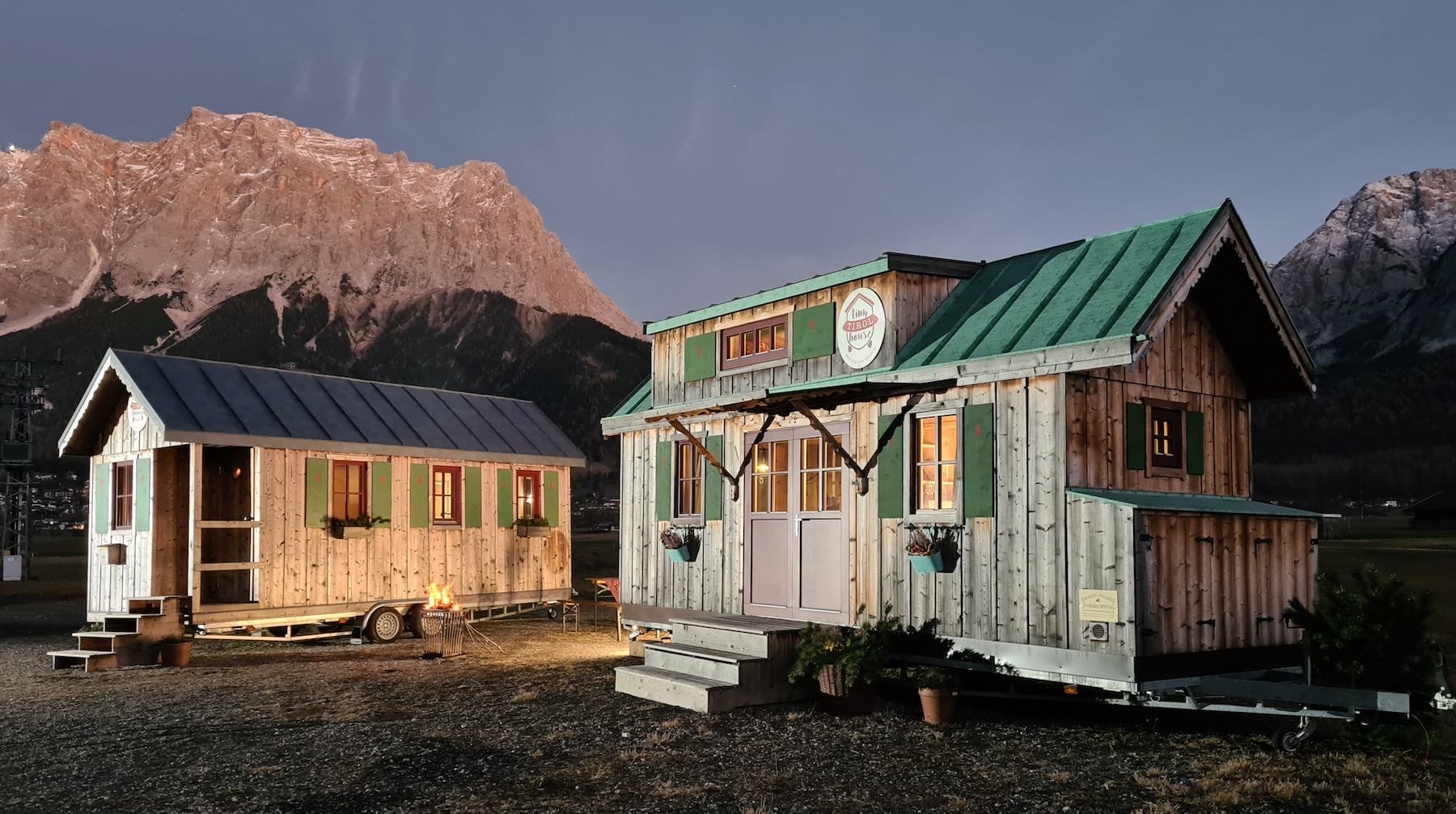 Zwei Tiny Houses bei Dämmerung vor einem beleuchteten Berg. Vor dem linken Haus brennt ein Feuer