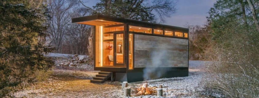 Modernes Tiny House mit Holzfassade und heller Beleuchtung in verschneiter Waldlandschaft, davor ein Lagerfeuer