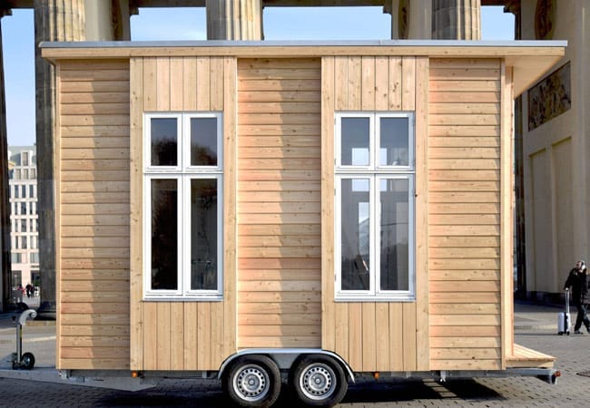 Ein Tiny House aus Holz mit weißen Fenstern, vor dem Brandenburger Tor in Berlin