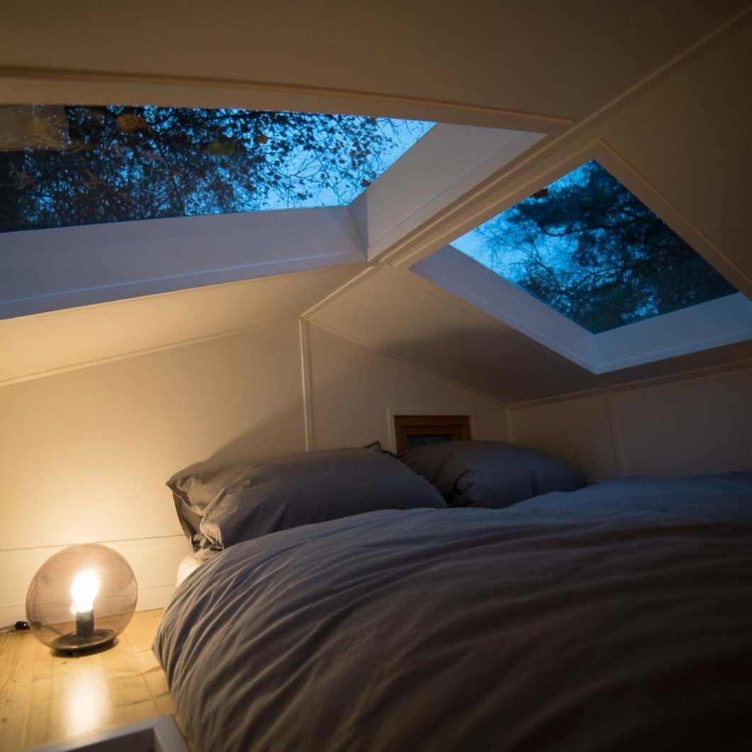 Bett mit grauer Bettwäsche und Kissen unter Dachfenstern mit Blick auf Baumkronen bei Dämmerung, schwaches Licht