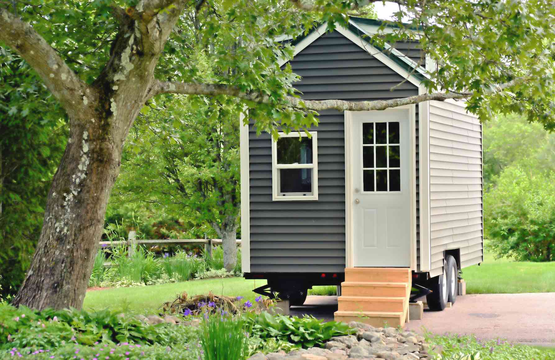 Ein Tiny House mit dunkelgrauer Fassade, weißer Eingangstür und kleinen Holzstufen im Grünen