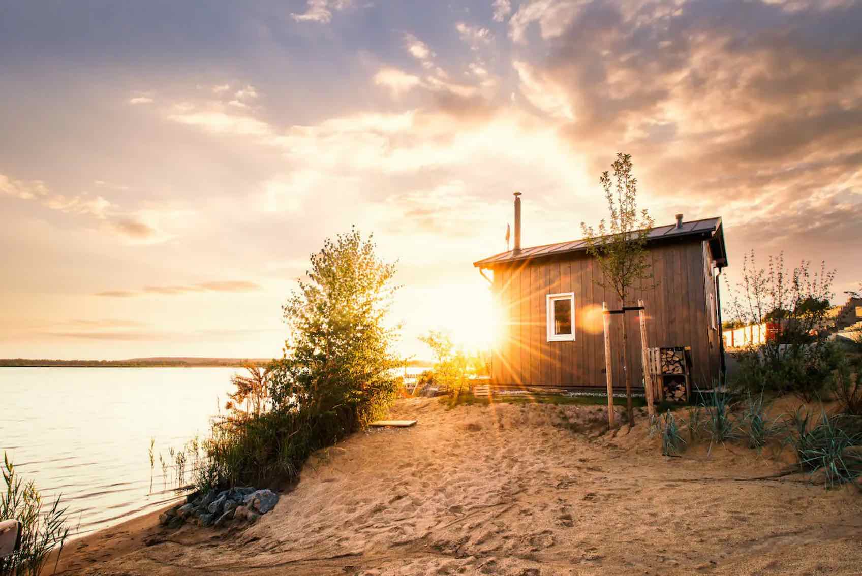 Sonnenuntergang am See, ein Tiny House aus Holz steht am Ufer auf Sand, neben Bäumen und Schilf