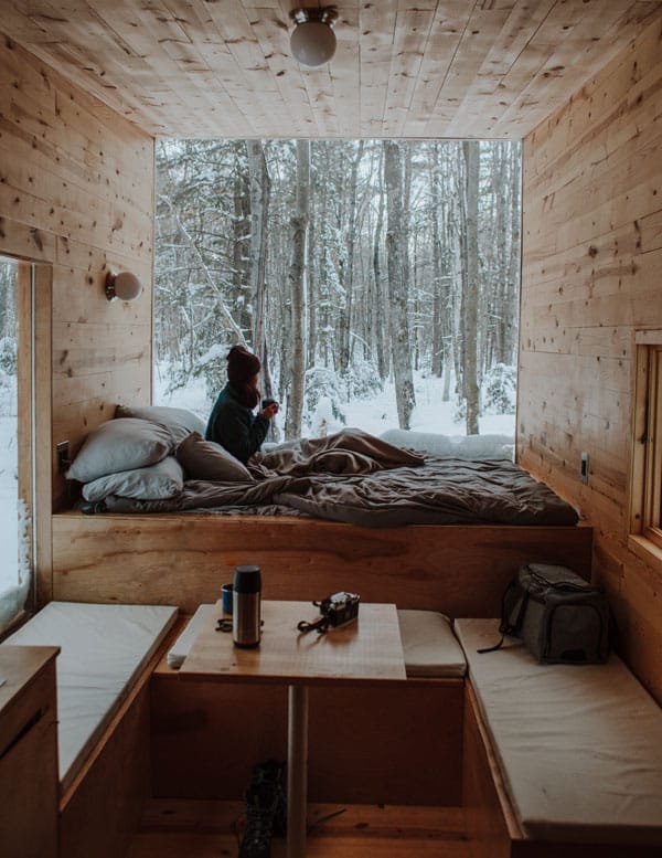 Eine Person sitzt in einem Tiny House und blickt aus einem großen Fenster in eine verschneite Waldlandschaft